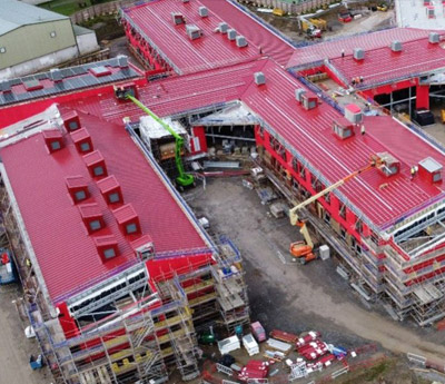 Aerial drone view of the Mynydd Isa campus under construction, showcasing the bright red metal roofing and facade of the multi-wing educational facility. The image captures the Robertson Construction site, the Wepco delivery partner, featuring extensive scaffolding, cherry pickers, and organised construction materials during the building phase.