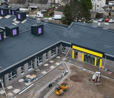 High-angle aerial view of a modern L-shaped commercial building under construction with a dark grey standing seam metal roof and prominent skylights. The site features grey brickwork, yellow architectural accents, and active construction equipment including an excavator and dumper truck on a dirt ground.