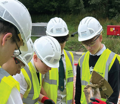 A group of students in school uniforms wearing Morgan Sindall Construction white hard hats and high-visibility vests participate in a hands-on social value workshop. The image shows the children gathered around a construction professional, engaging in a practical building exercise on-site as part of an educational career outreach program.