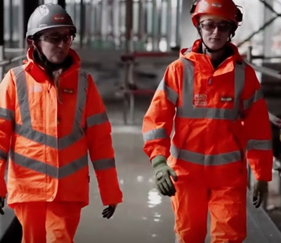 A medium full, slightly low angle photo shows two women construction workers walking toward the camera on a platform. They both wear bright orange hardhats, matching work coveralls with reflective stripes, work gloves, and sunglasses. The woman on the right has ear protection over her ears. The background is a maze of metal scaffolding and beams, with light streaming from open windows in the background.